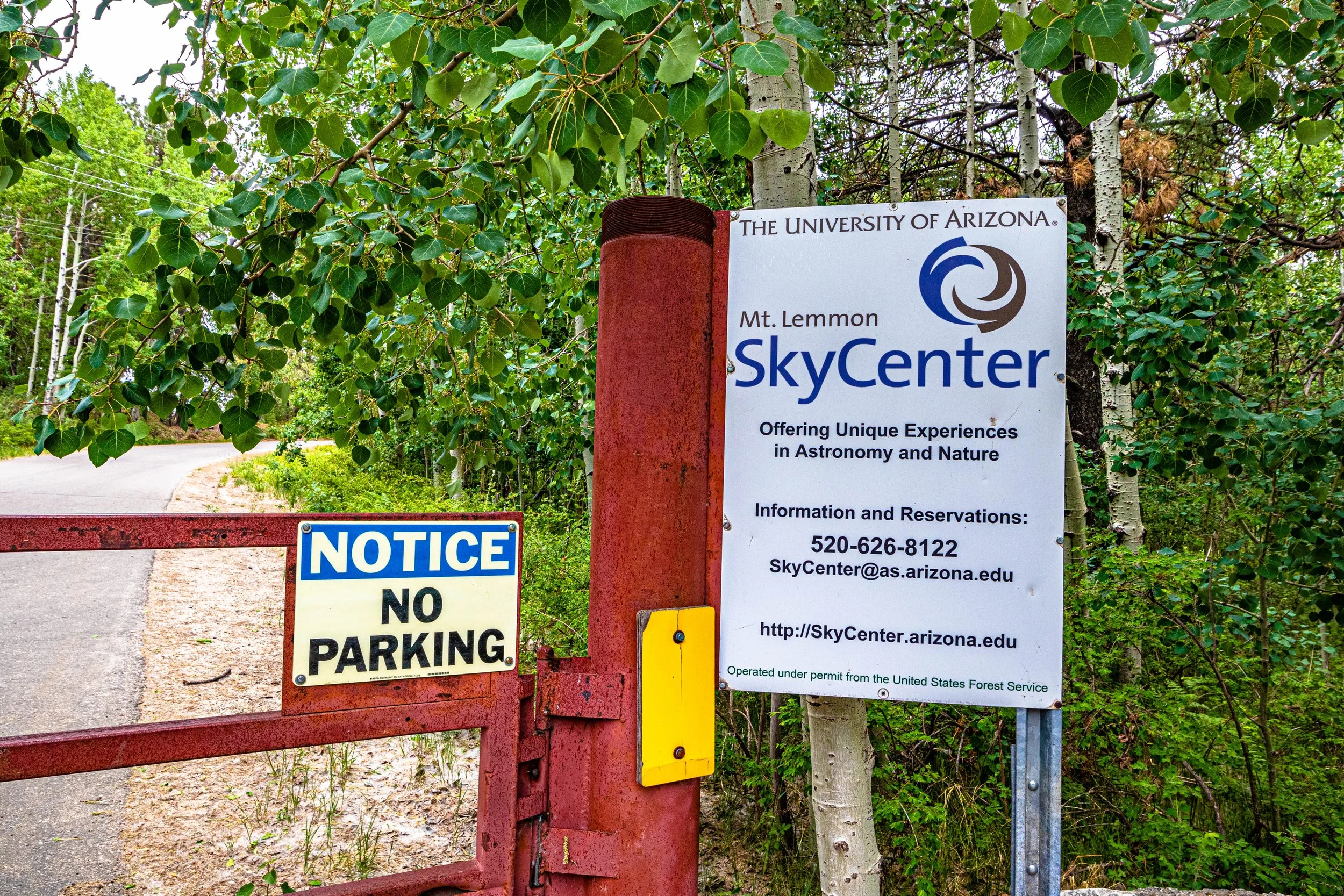University of Arizona Mount Lemmon SkyCenter sign beside a red entrance gate surrounded by green trees.