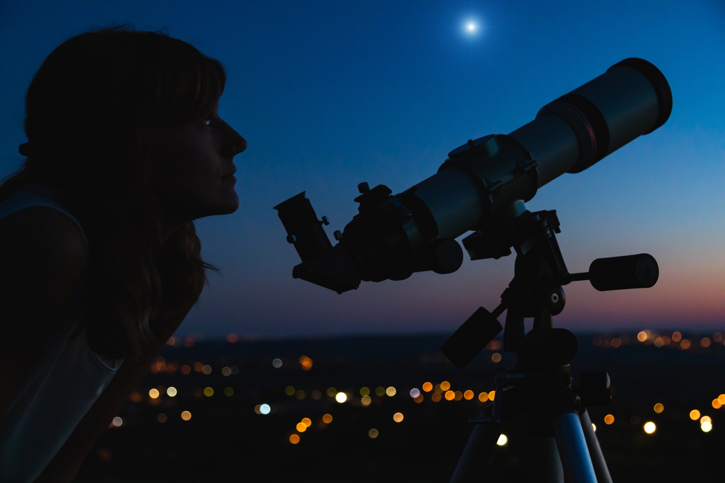 Woman beside a telescope at twilight, with city lights glowing in the background.