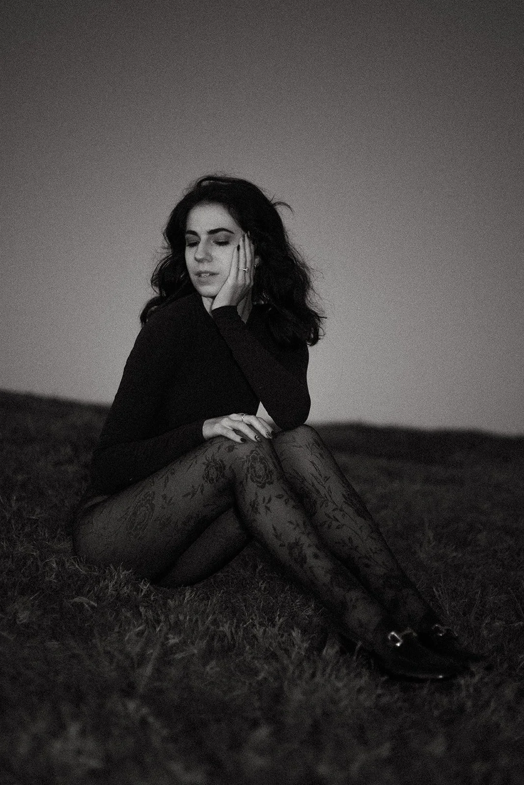 brunette woman sitting on a grassy hill. the photo is in black and white. the woman is wearing a black long sleeved shirt with black floral tights.