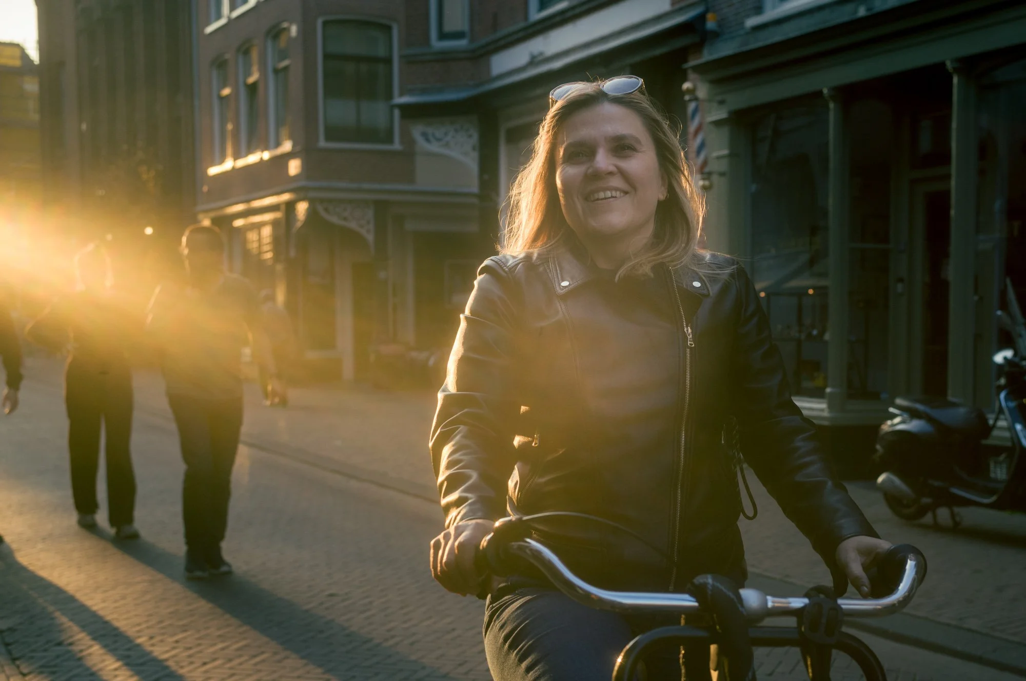 A woman riding a bicycle on a city street during sunset, with sunlight shining behind her and other pedestrians in the background.