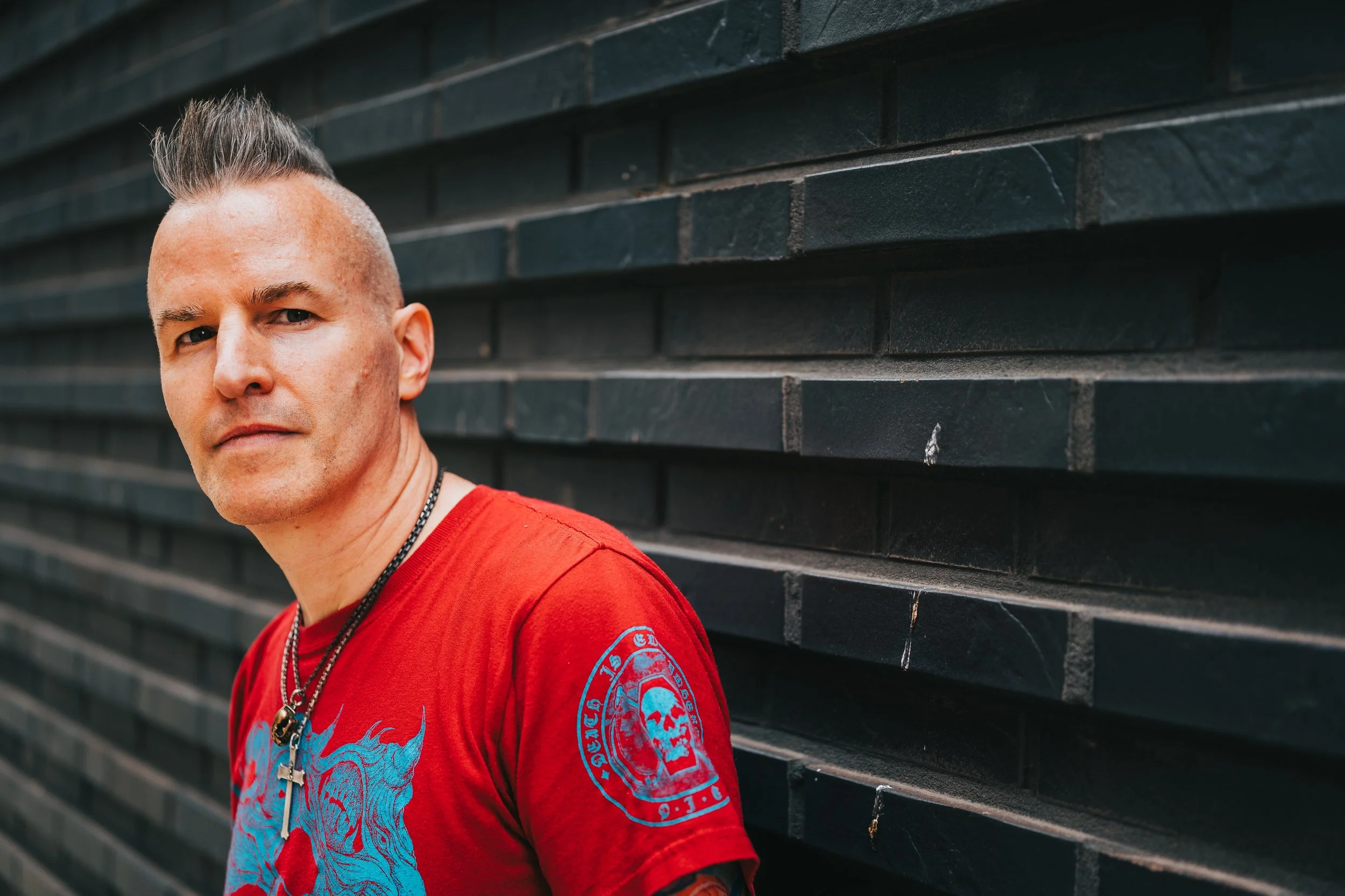 A man with a mohawk hairstyle wearing a red shirt and necklaces standing next to a black brick wall.