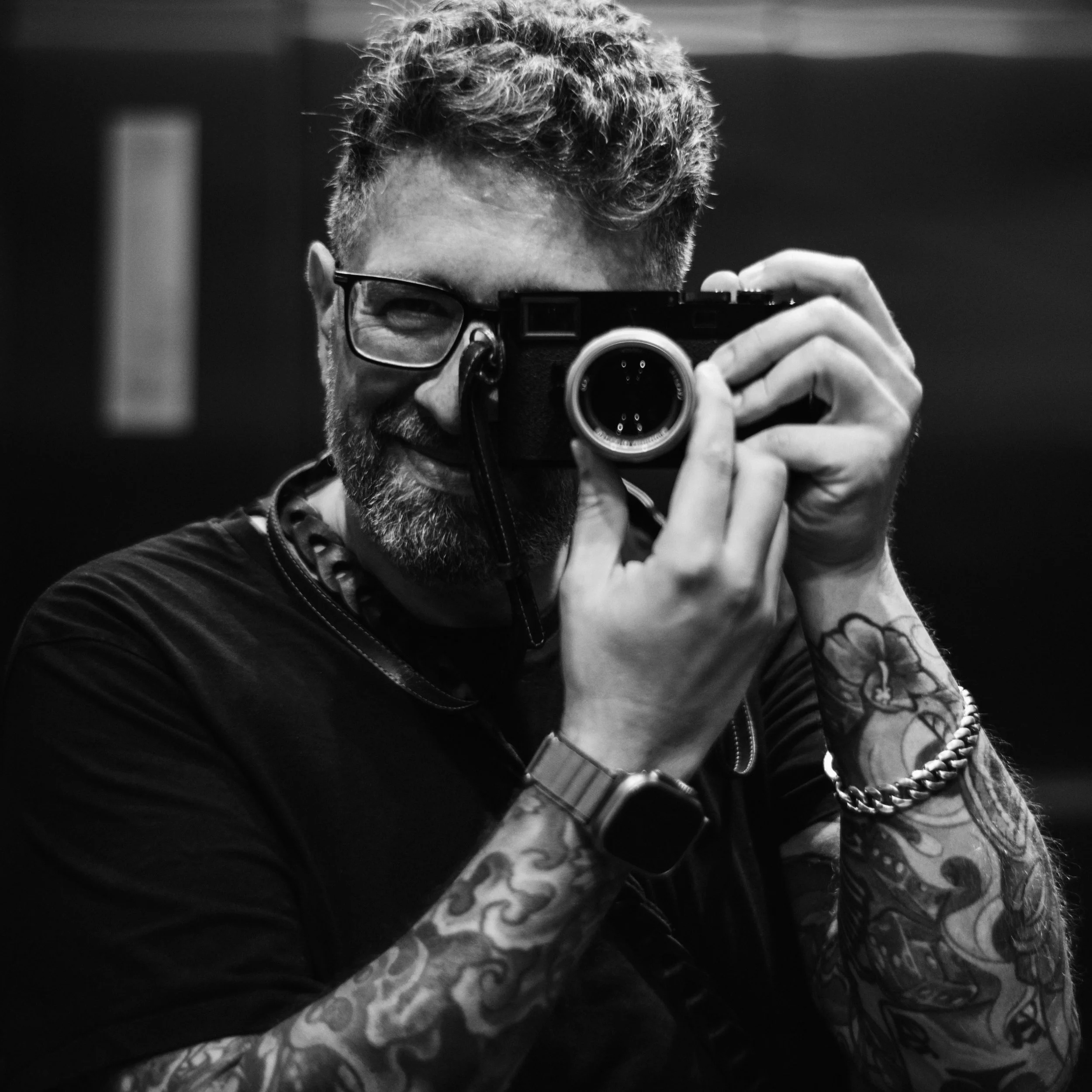A man with glasses and a beard taking a mirror selfie with a camera. He has tattoos on his arm and is wearing a dark shirt.