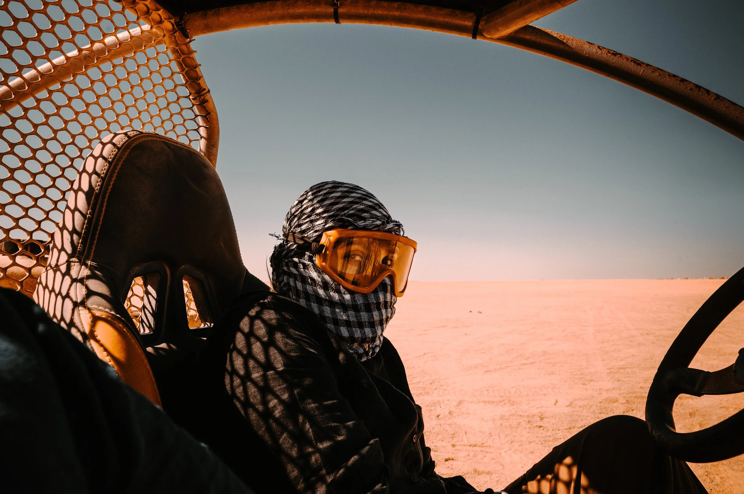 Two individuals sitting in a vehicle, wearing scarves and protective eyewear, with sand dunes and a clear sky visible in the background, suggesting a desert setting.