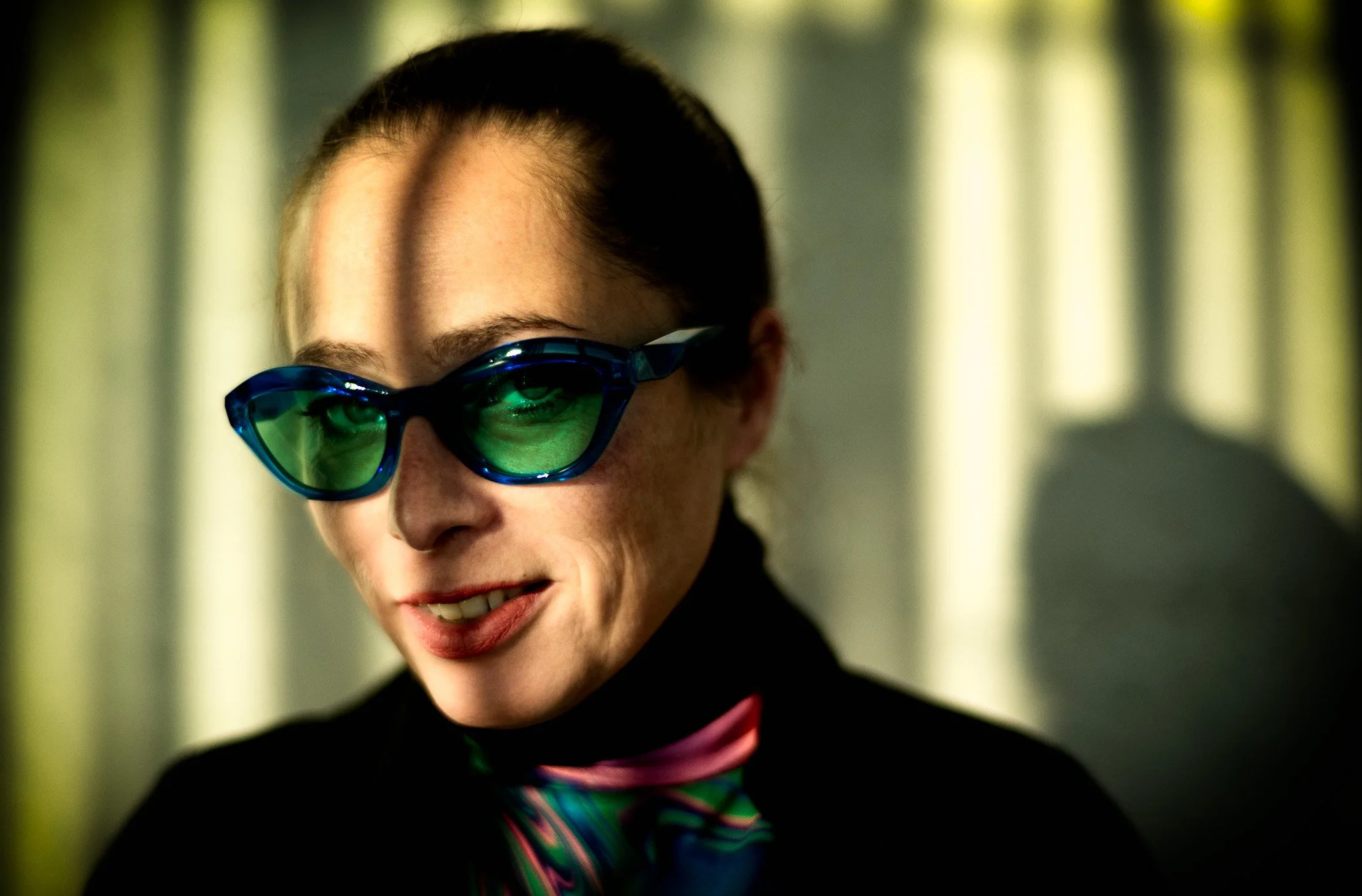 Close-up of a person wearing reflective sunglasses, smiling, with dark hair pulled back, and wearing a colorful, patterned scarf or tie.