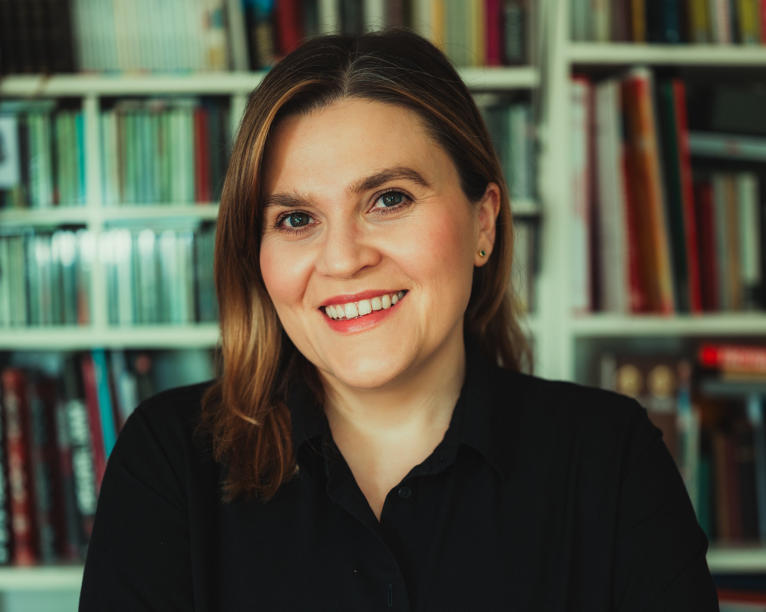A woman with brown hair and blue eyes smiling in front of a bookshelf filled with colorful books.