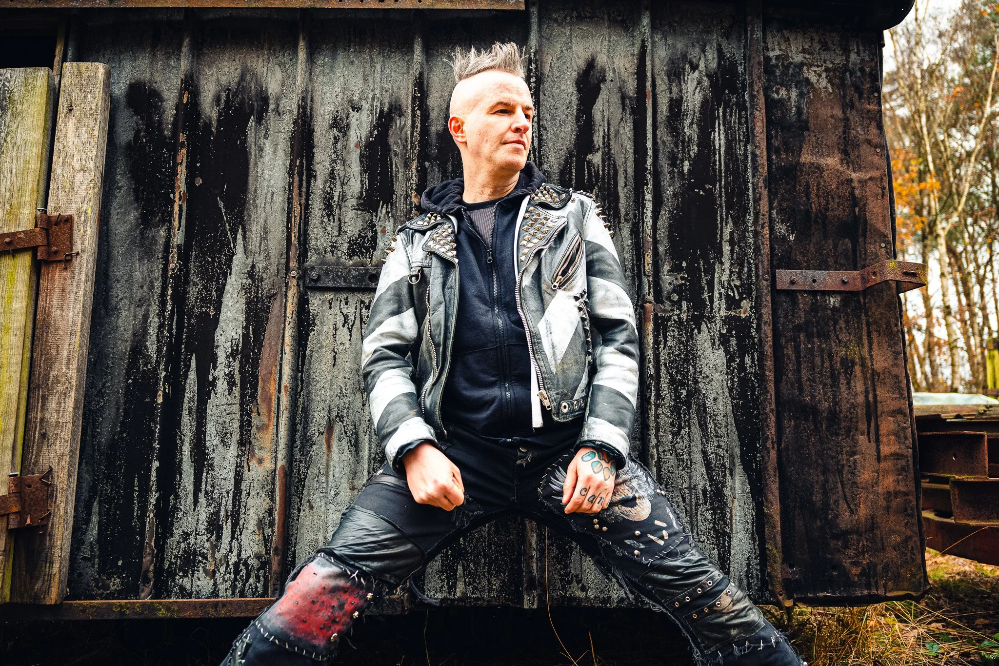A man with a shaved and spiked hairstyle, wearing a studded black and gray leather jacket, black shirt, and distressed leather pants with red and black paint marks, standing in front of a weathered black wooden shed with rusted metal hinges. There ar