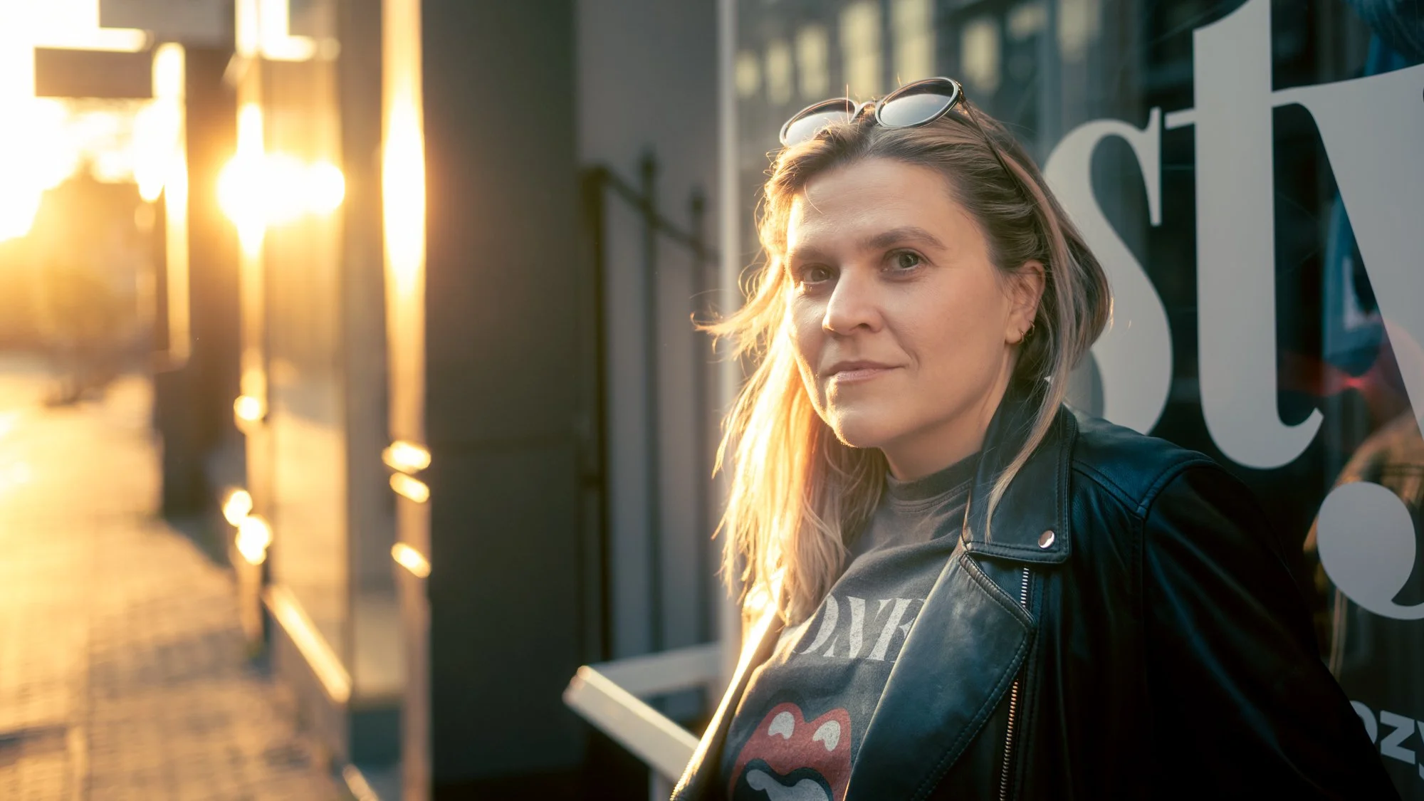 A woman with blond hair wearing sunglasses on her head and a black leather jacket, standing outside during sunset, leaning against a window with white text.
