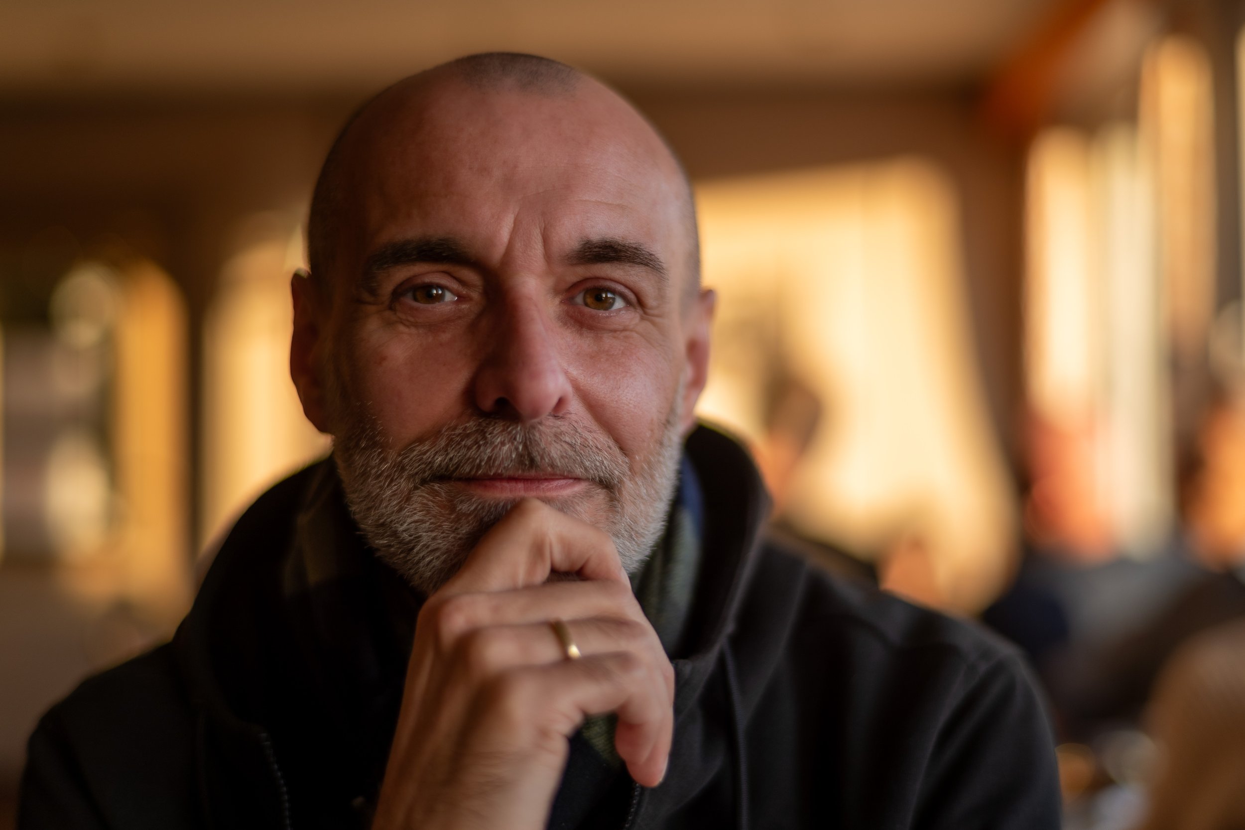Close-up of an older man with a beard, bald head, and brown eyes, looking into the camera and resting his chin on his hand, in a warmly lit indoor setting.