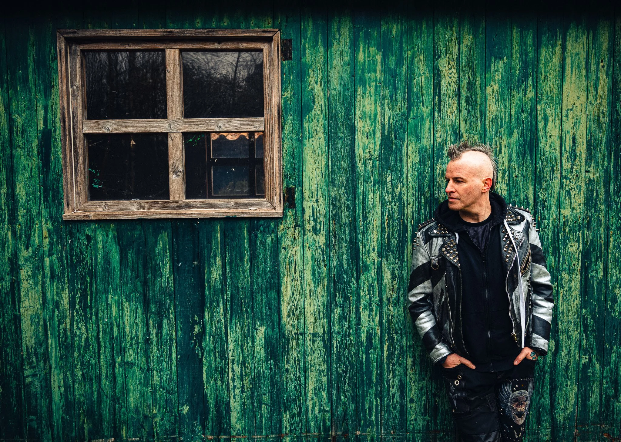 A man with a mohawk hairstyle and a punk leather jacket standing against a weathered green wooden wall with a small window. The man has a serious expression and is looking to the side.