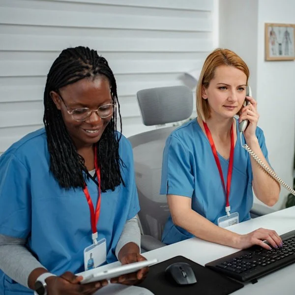Two female healthcare workers in blue scrubs working at a desk, one looking at a tablet and the other on the phone.