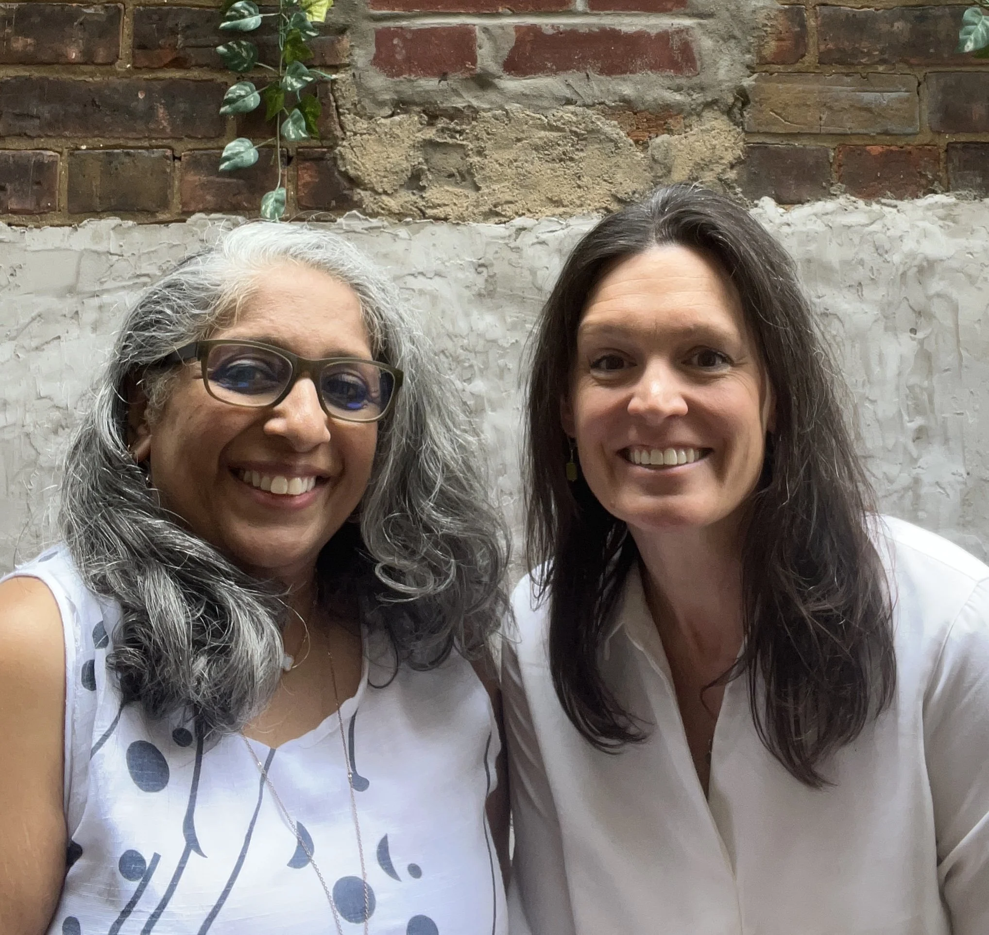 Two women smiling for a photo in front of a brick wall. One woman has gray hair, glasses, and headphones, and the other has dark hair.