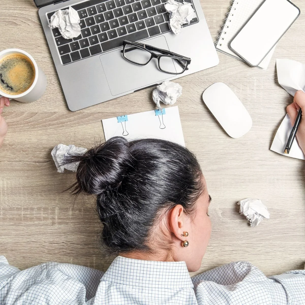 A person with dark hair tied in a bun sleeping at a cluttered desk with crumpled papers, a laptop, a coffee mug, a pair of glasses, a notepad, a pen, a paper with paper clips, and a mouse.