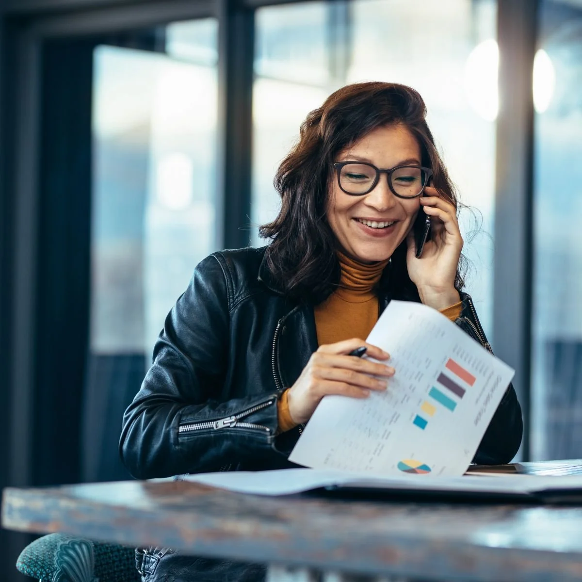 A woman with glasses and dark wavy hair smiling while talking on her cellphone and looking at a printed graph with colorful charts.