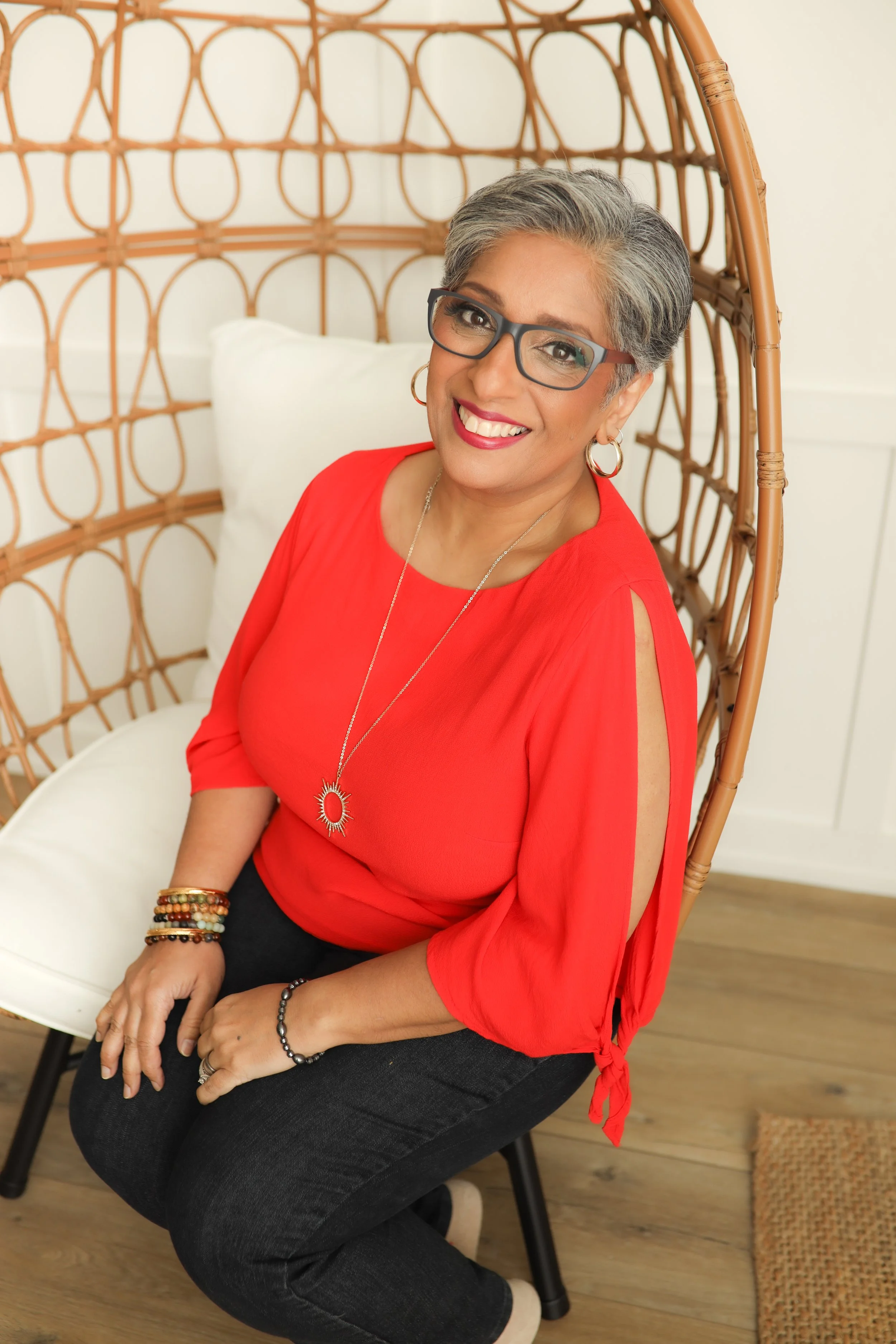 A smiling woman with short gray hair and glasses, wearing a red top, sitting on a black chair with wooden legs. She is accessorized with jewelry including bracelets, earrings, a necklace, and a ring. The background features a white wall and a rattan hanging chair with a white cushion, on a wooden floor.