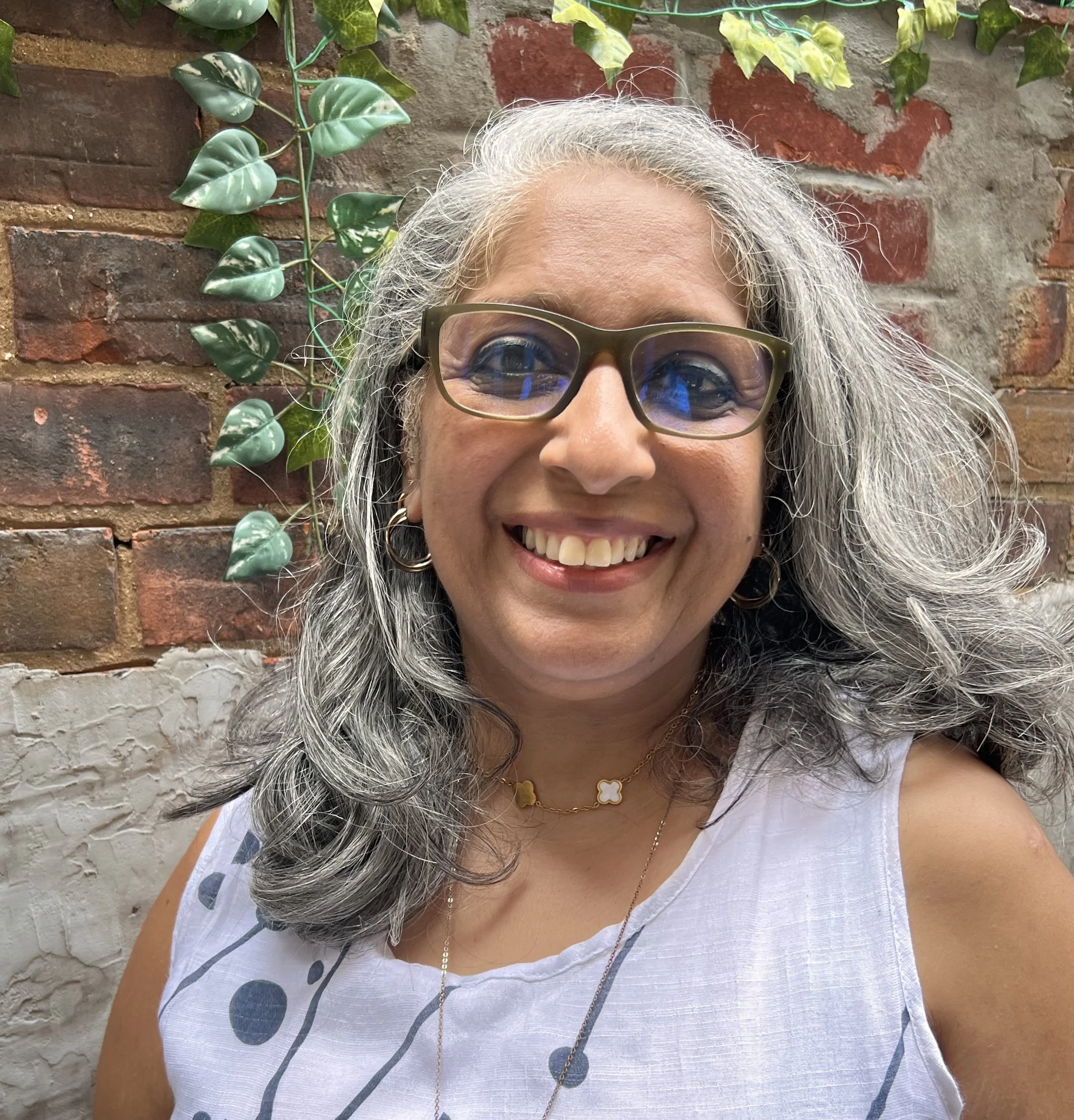 Close-up of an older woman with grey curly hair, glasses, smiling, standing against a brick wall with green ivy plants.