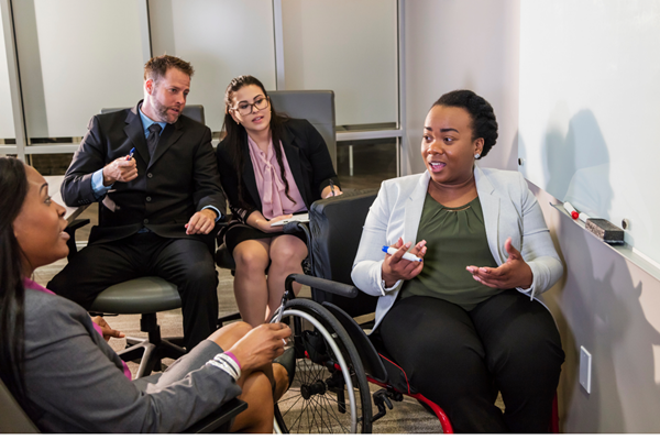 Four diverse business professionals engaged in a discussion in a conference room, with one woman in a wheelchair speaking and the others listening attentively.
