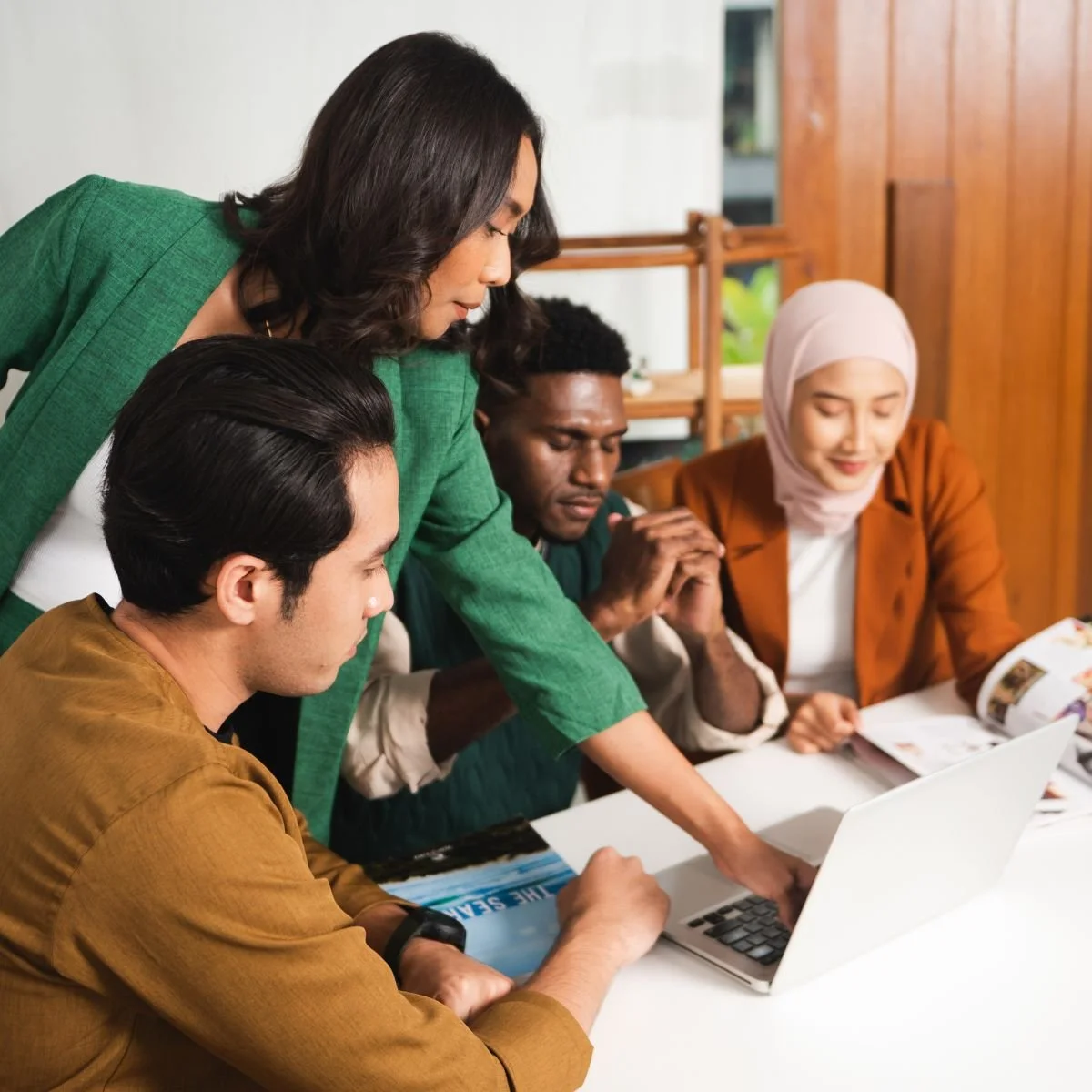 A diverse group of four people gathered around a table looking at a laptop, with one woman standing and pointing at the screen, as they work together in a bright, modern office or cafe setting.