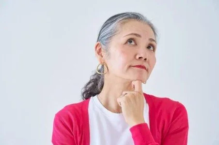 An older woman with gray hair tied back, wearing a white shirt and red cardigan, looking upward thoughtfully with her hand on her chin against a plain white background.