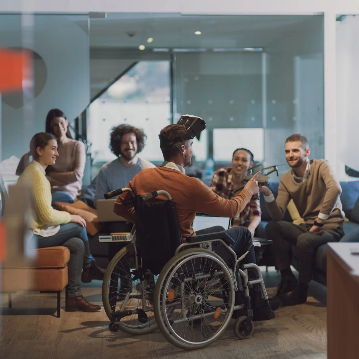 Group of five diverse people sitting and laughing in a modern office lounge, with one person in wheelchair wearing a virtual reality headset, pointing at and engaging with others, some holding laptops.