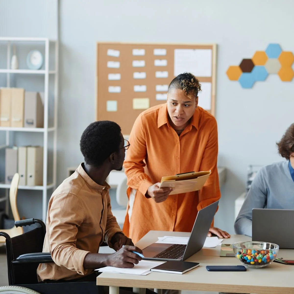 A diverse group of people working together in an office, with one woman standing and showing something on a tablet to a person in a wheelchair.