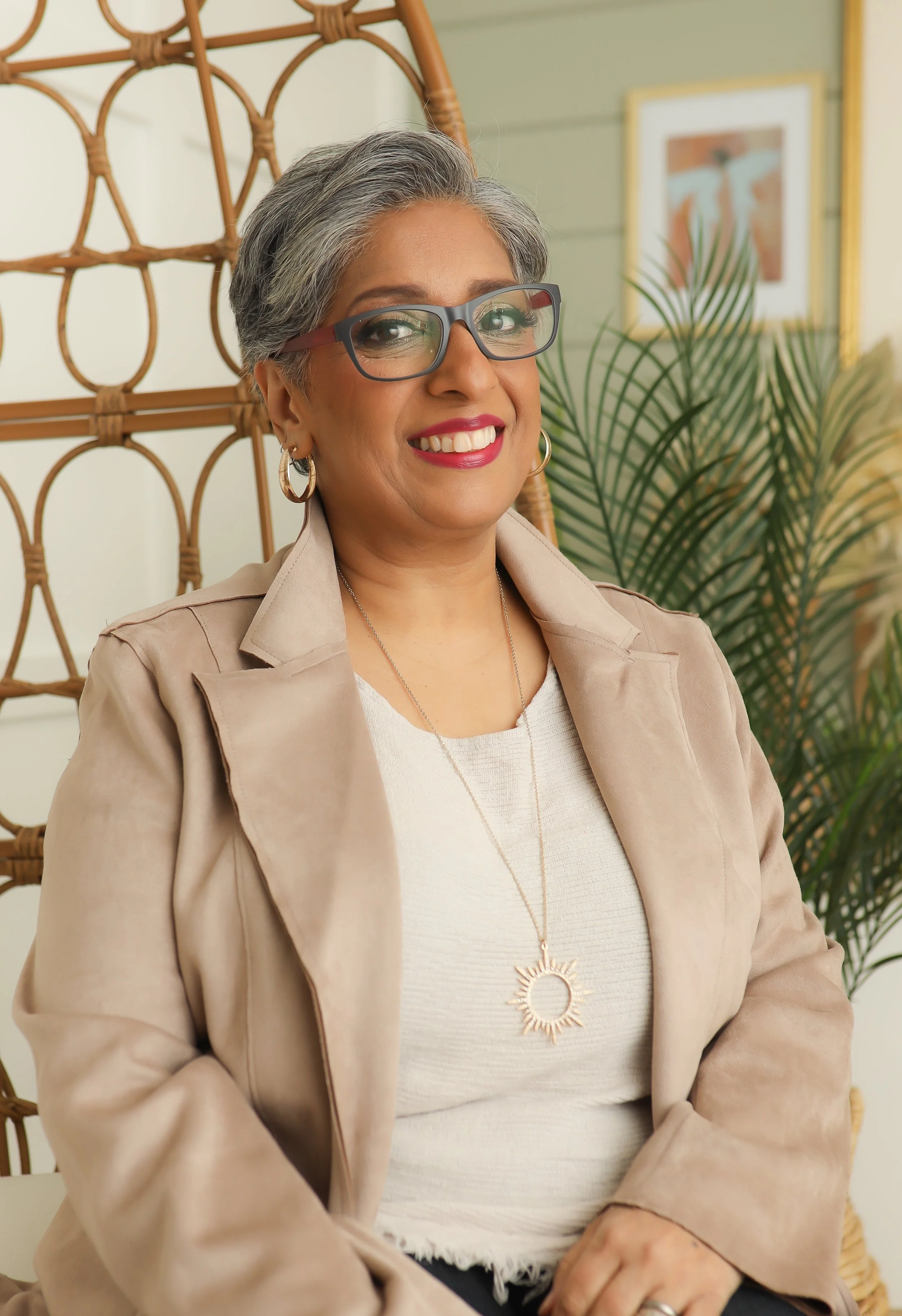A smiling woman with gray hair, wearing glasses, earrings, a beige jacket, and a white top, sitting indoors with a large green plant and framed artwork in the background.