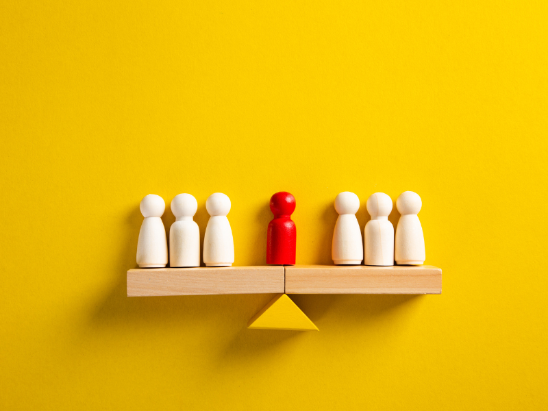 Wooden balance scale with five white figurines on the left, one red figurine in the center, and three white figurines on the right, set against a yellow background.