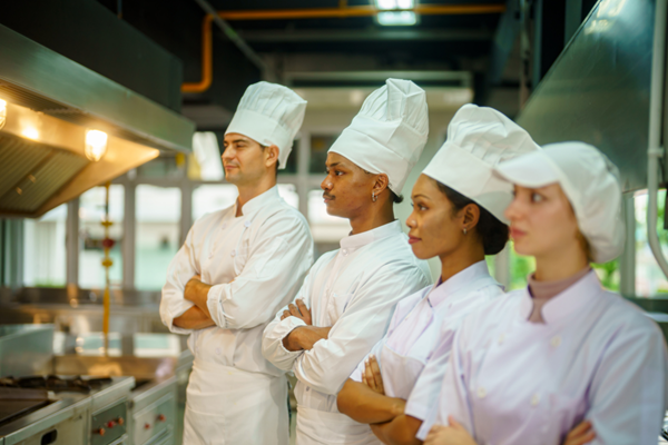 Four professional chefs wearing white uniforms and tall white hats stand in a commercial kitchen, observing a cooking process.