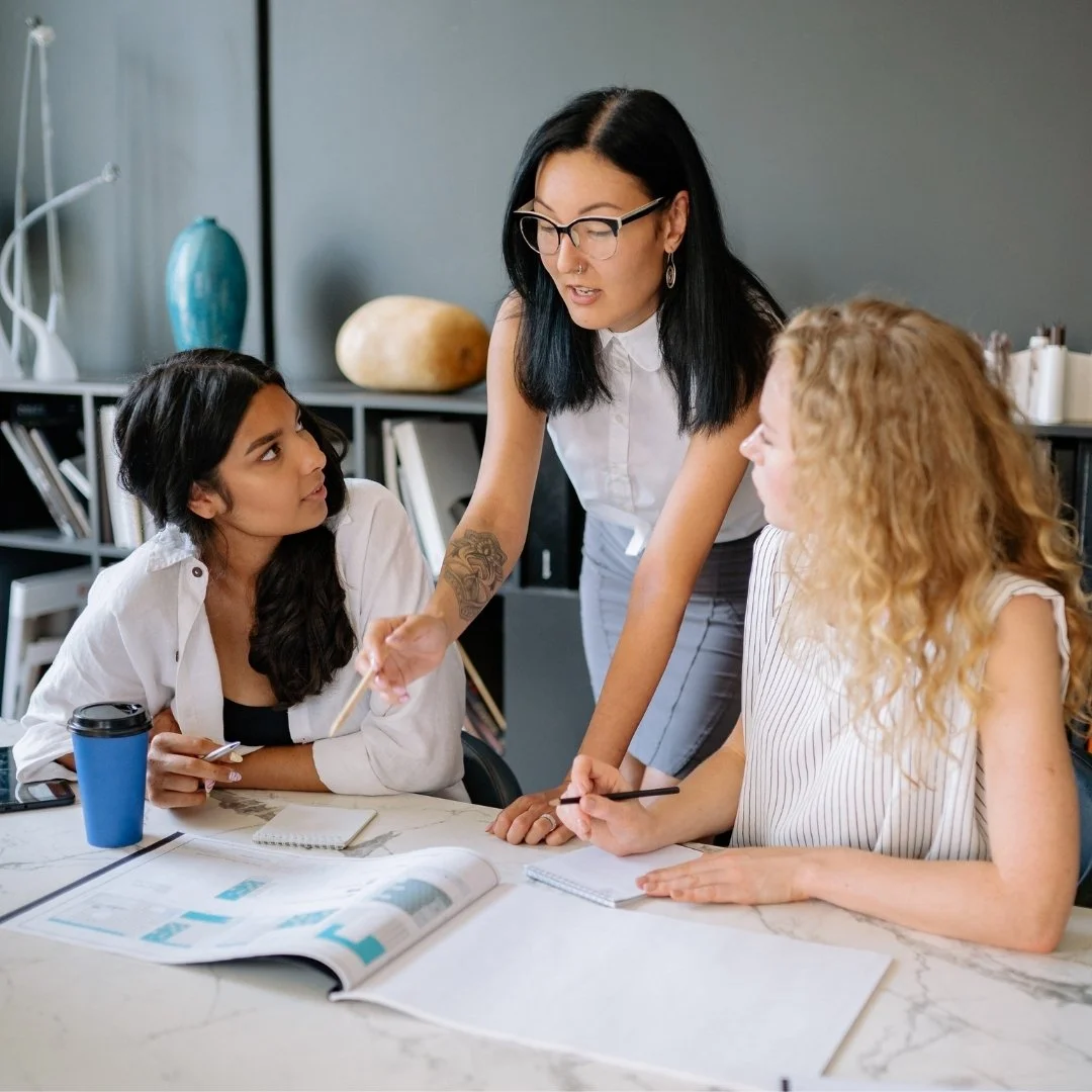 Three women having a discussion at a table in an office setting, with notebooks, papers, and a coffee cup in view.