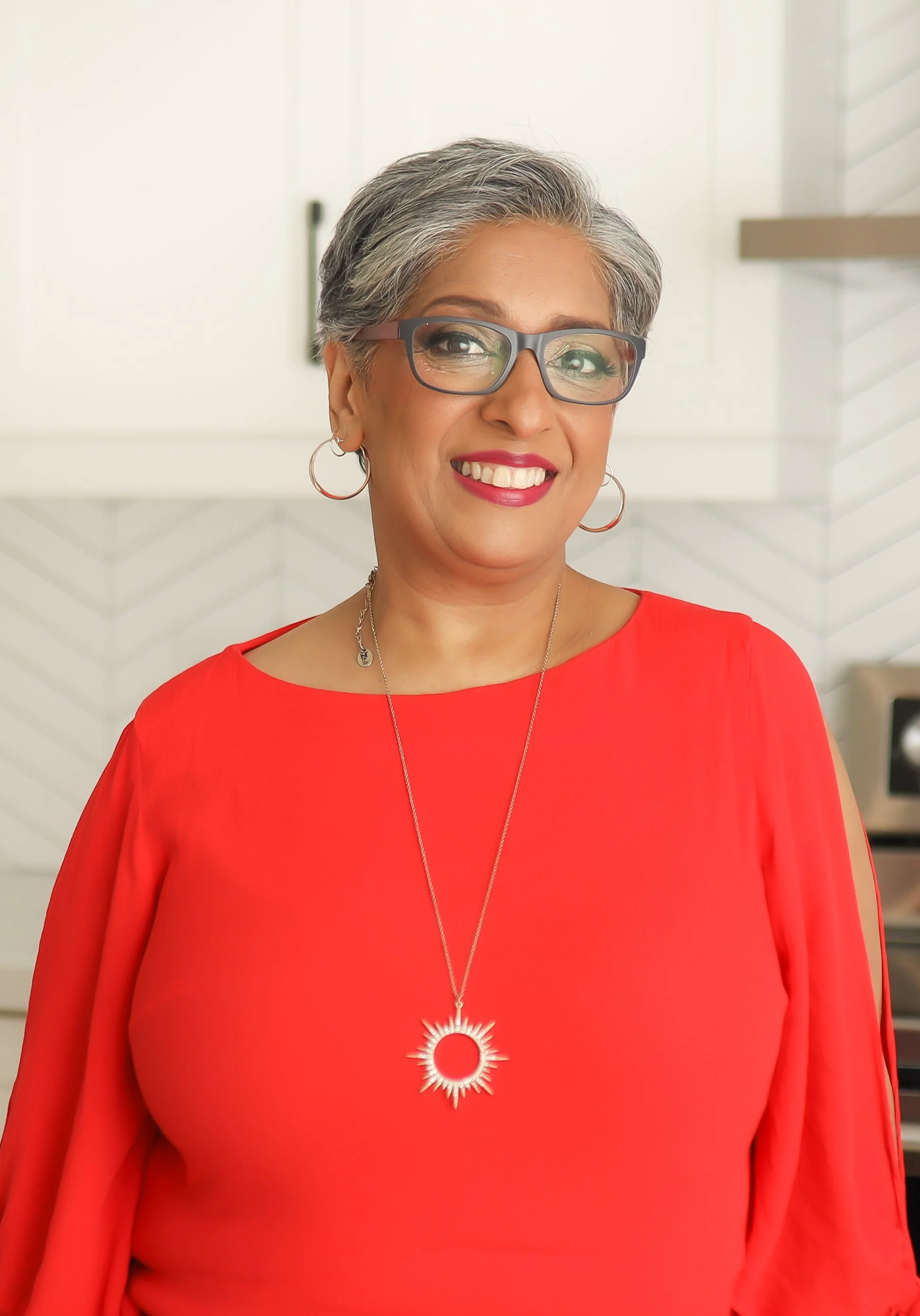 A woman with short gray hair and glasses, wearing a bright red dress and a sun-shaped pendant necklace, smiling in a modern indoor setting.