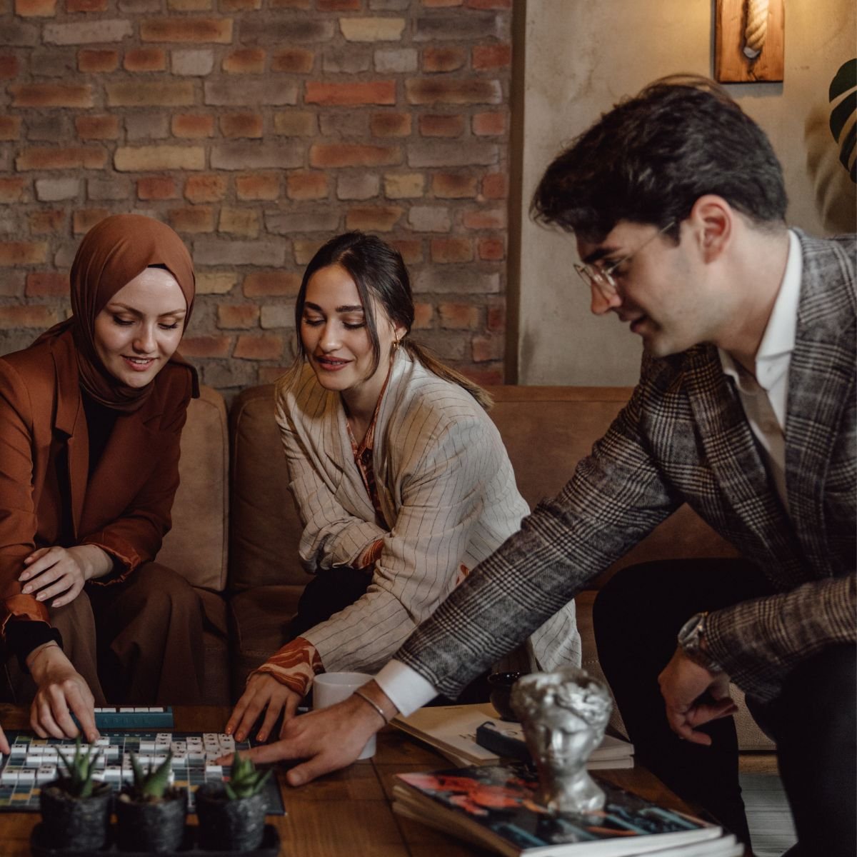 Three people sitting around a table playing a board game and smiling, in a cozy room with an exposed brick wall.
