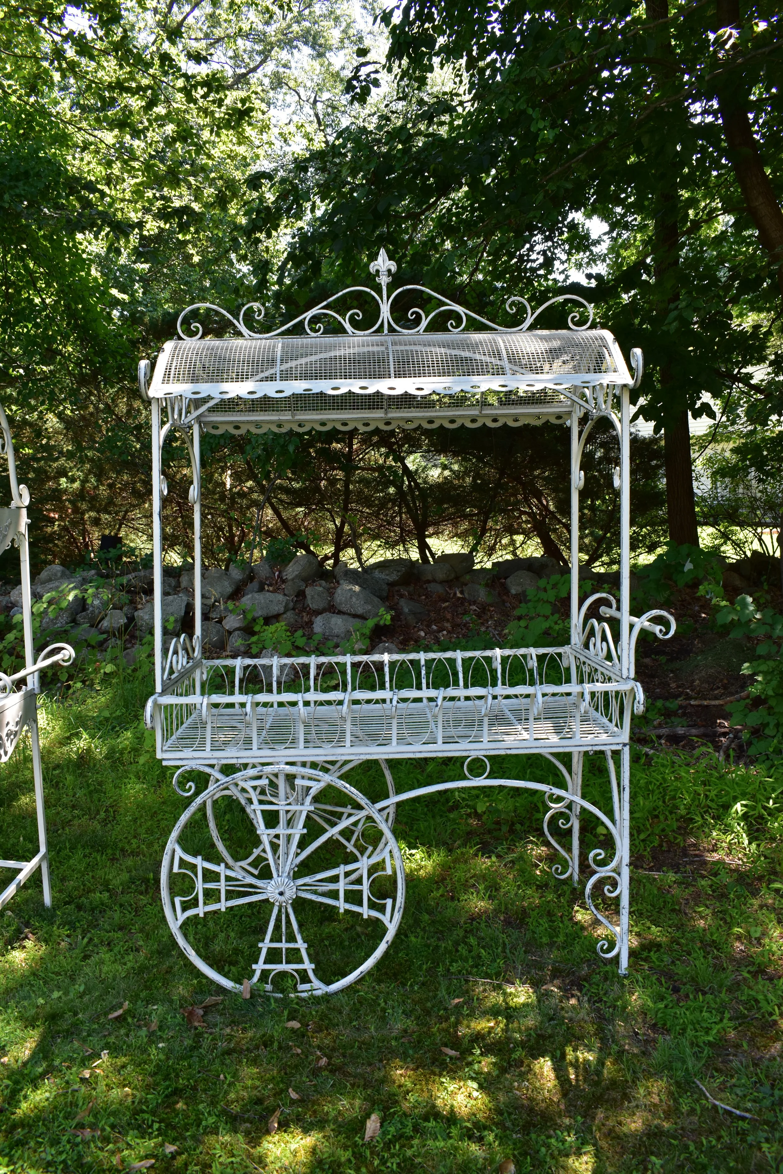 An ornate white metal cart with a floral arrangement on top, including pink, white, and purple flowers, and greenery. There are directional signs attached to the cart with labels like 'Tea Party,' 'Turn Around,' 'Wonderland,' 'This Way,' 'That Way,' and 'Wrong Way.' 