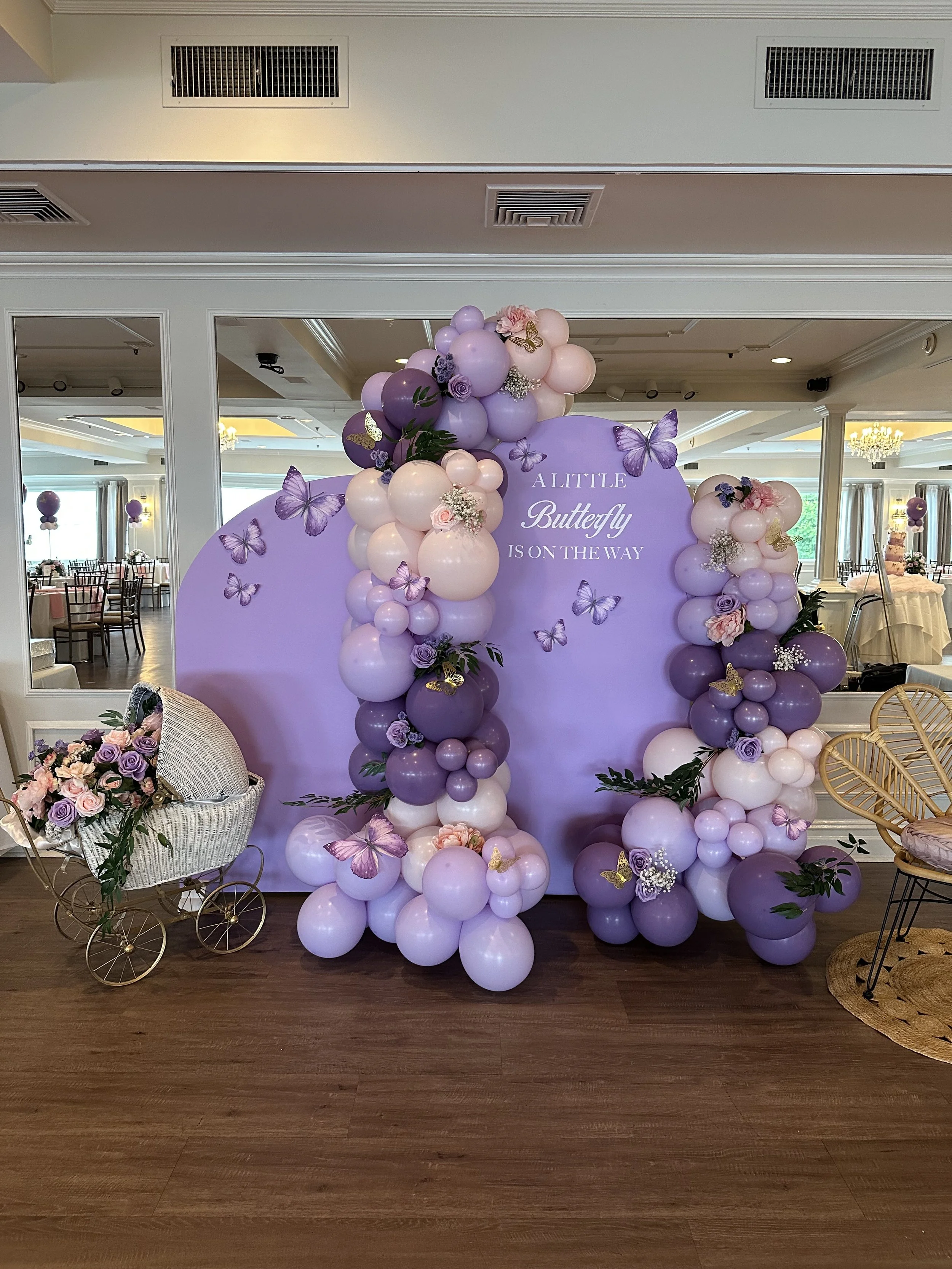 Decorative balloon display in shades of purple, pink, and white with flowers, butterflies, and a sign reading 'A little butterfly is on the way' at an event venue.