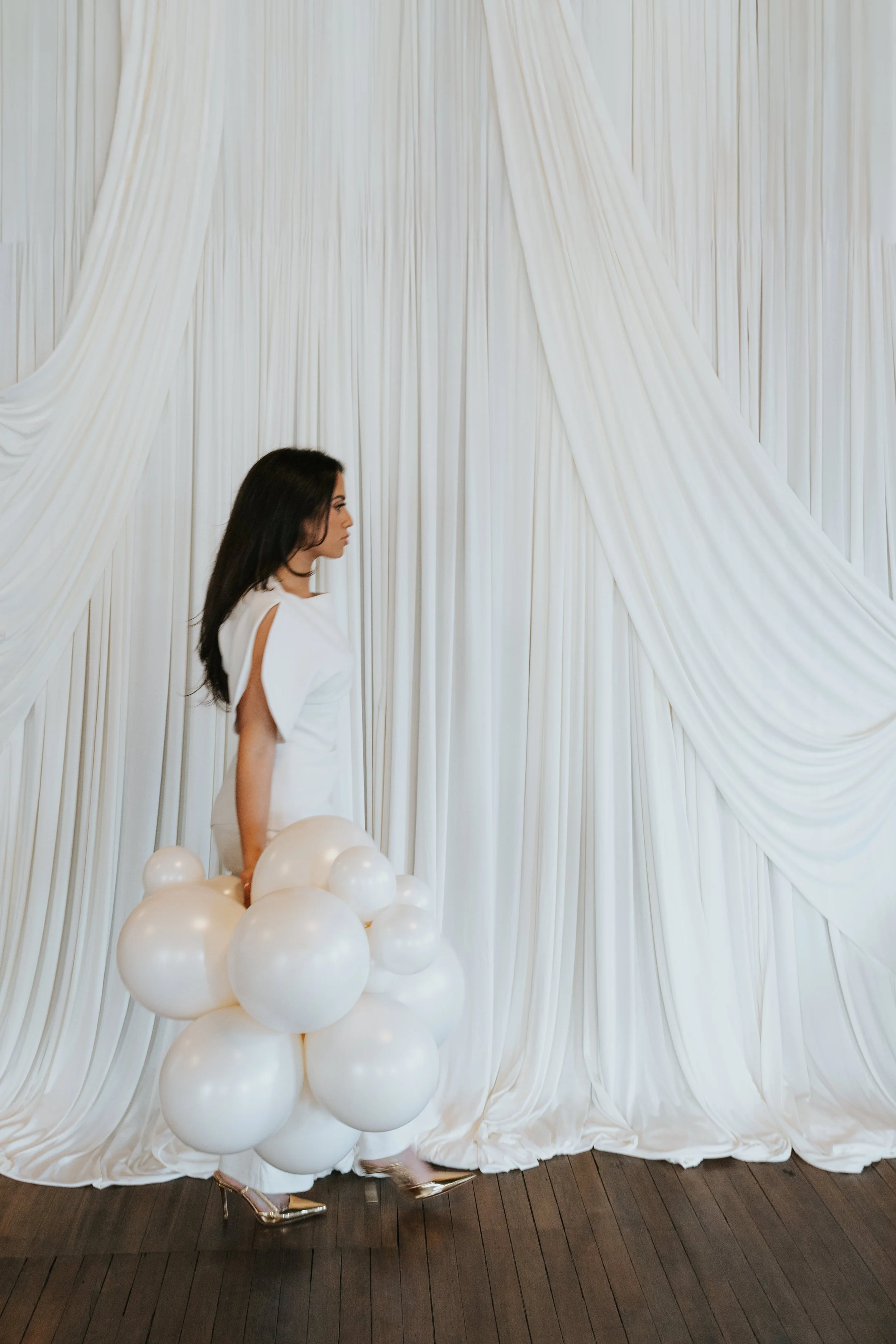 A woman in a white dress standing among white balloons with a background of cream-colored drapes.