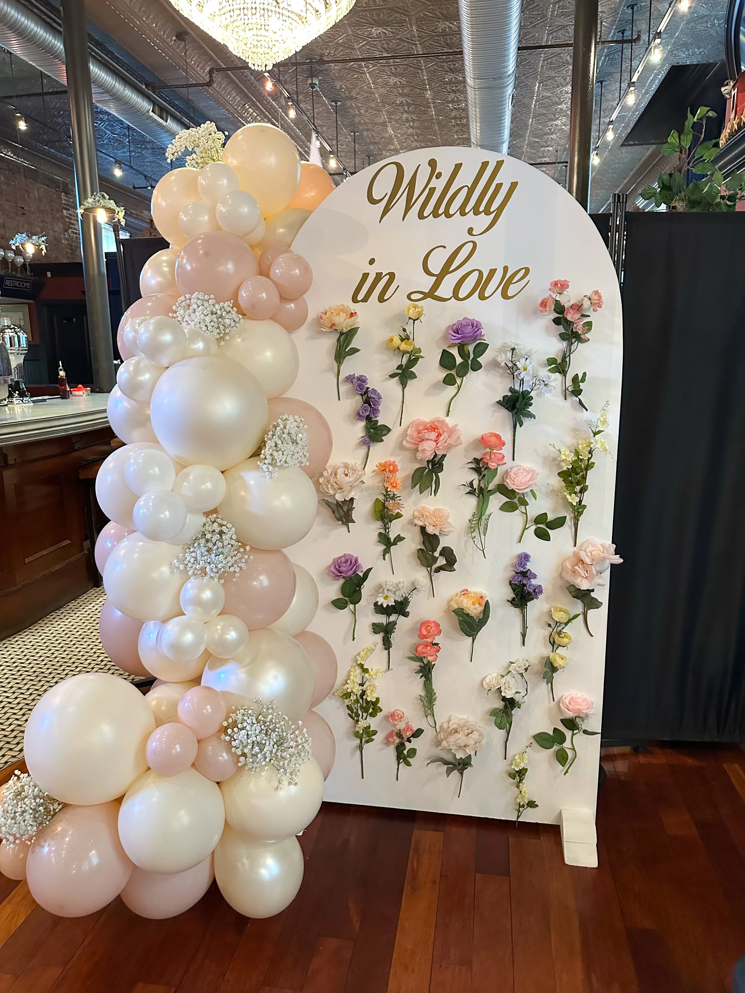 Decorative display Chiara panel that says 'Wildly in Love' in gold lettering, surrounded by pastel-colored balloons and flowers.