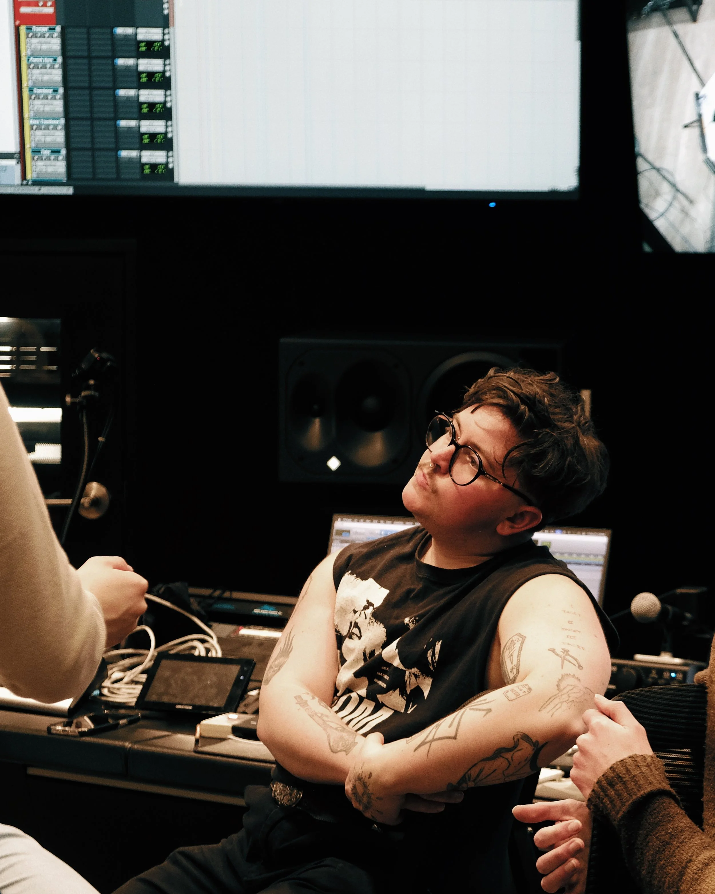 A person with tattoos on their arms, wearing glasses and a black sleeveless shirt, is sitting in a recording studio with audio equipment and a computer screen in the background. Someone is handing them a device with multiple cords.