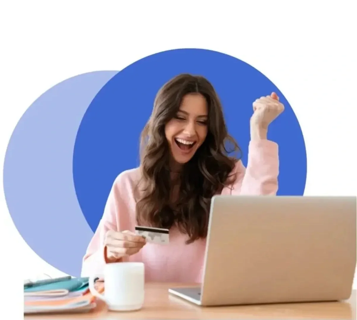 A woman sitting at a desk, holding a credit card in one hand and celebrating with a joyful expression, next to a laptop and a coffee mug.
