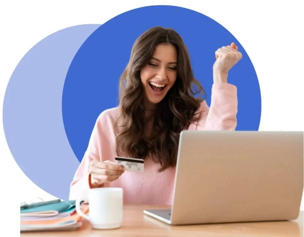 A woman celebrating while shopping online with a credit card, sitting at a desk with a laptop, mug, and notebooks.
