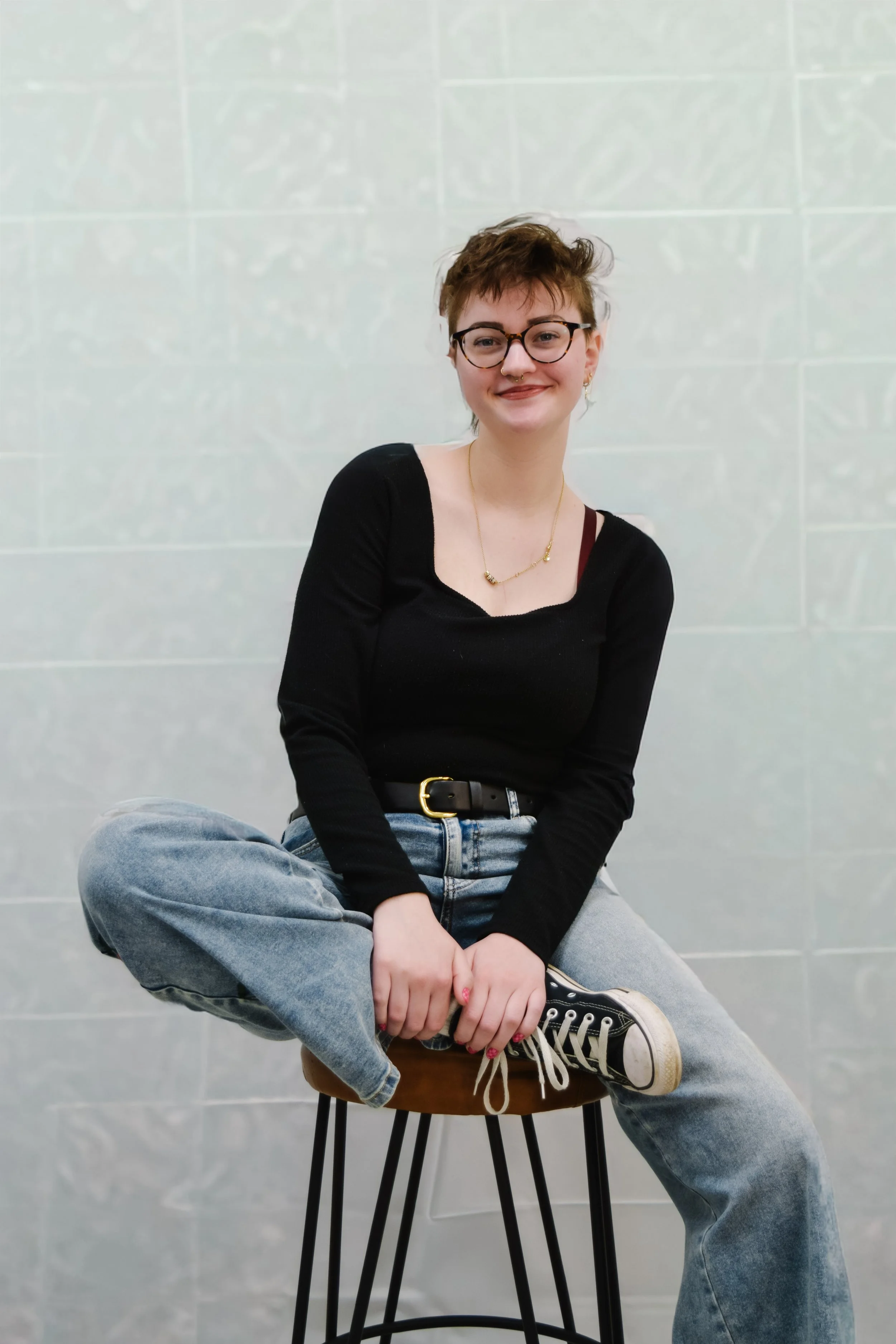 Sophia, nail technician at GROW Salon and Spa in Brookside Kansas City, smiling while seated on a stool wearing a black top and jeans