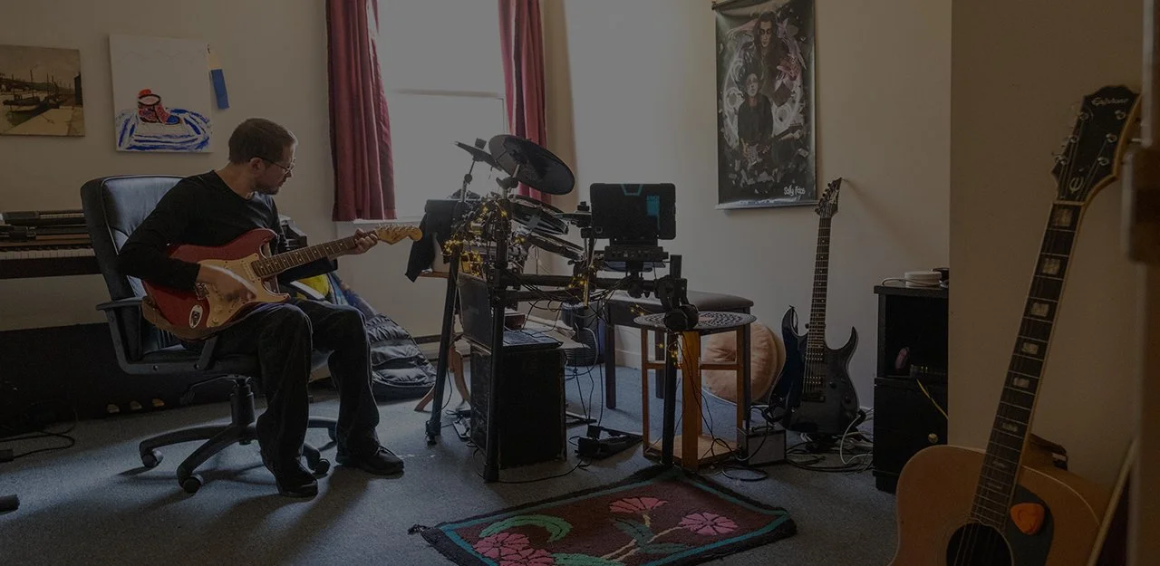 Man playing his guitar in his room with other musical instruments around him.