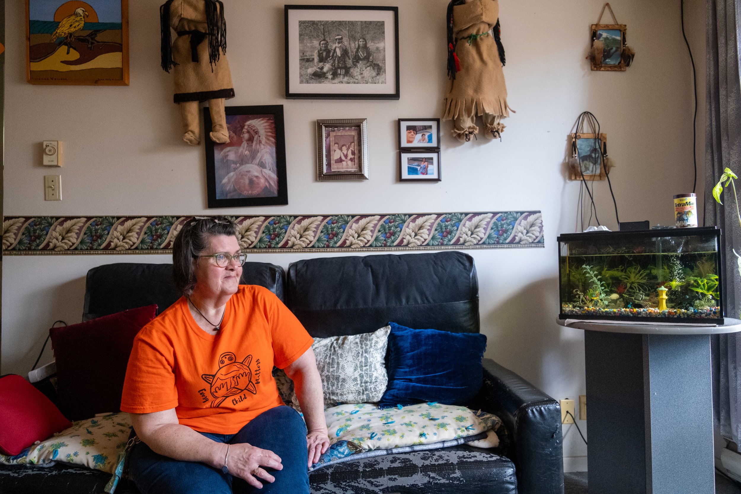 Indigenous woman in an orange t-shirt sitting on her couch. There is a fish aquarium to the side and many indigenous images on the walls.