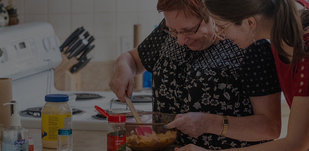 Woman making food in a kitchen while caretaker looks on.