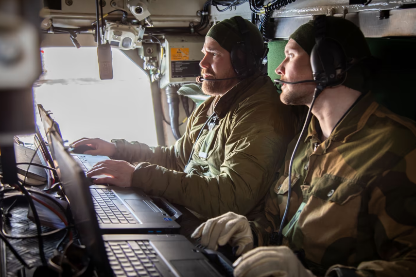 Two military personnel operating laptops inside a vehicle, wearing headsets and military uniforms.