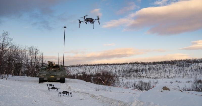 Military vehicle parked in snowy terrain with hovering swarm drones, surrounded by trees and a cloudy sky.