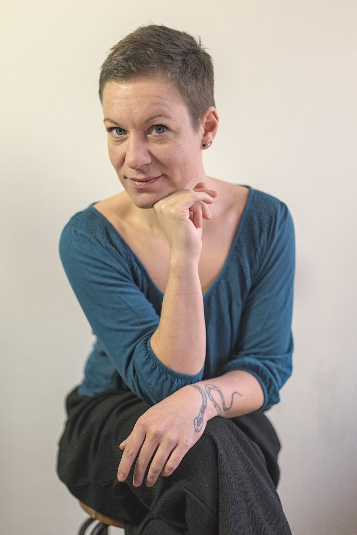 Sexual- und Beziehungscoach Eva Hanson mit türkisfarbenem Shirt vor weißem Hintergrund, sitzt auf einem Hocker und stützt das Kinn in die Hand