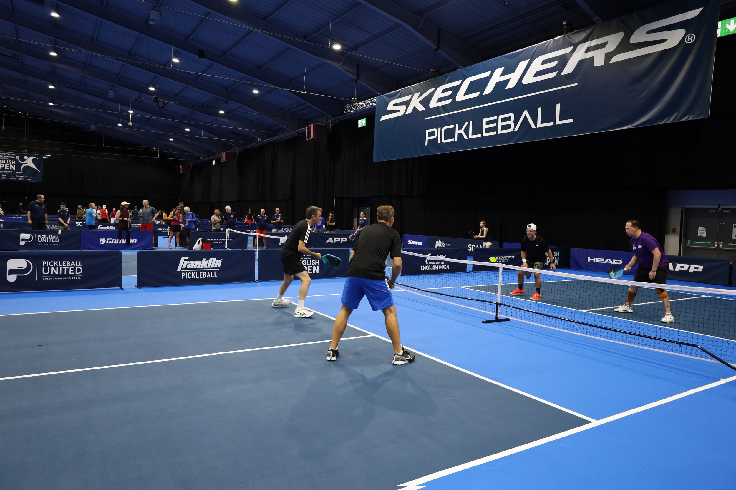 Indoors pickleball match with two teams of two players on a blue court, surrounded by advertisements for Skechers and Franklin Pickleball.