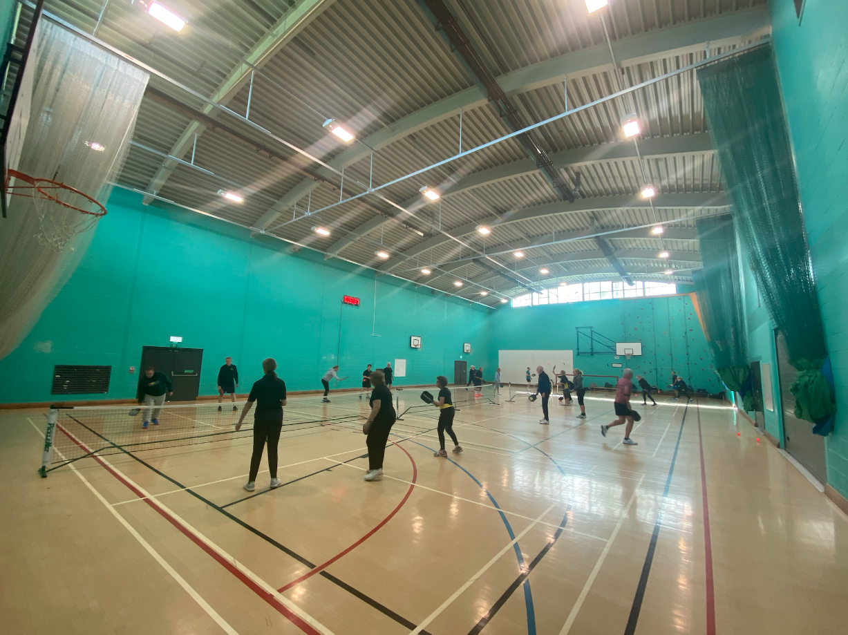 Indoor sports hall with people playing pickleball, marked courts, and teal walls.