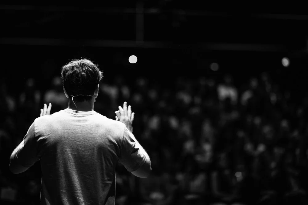 A person giving a presentation or speech in front of an audience, seen from behind in black and white.