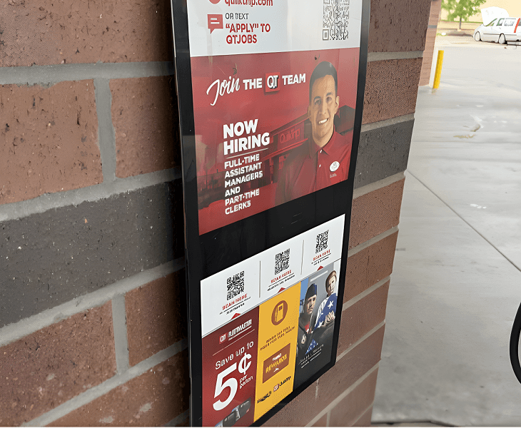 A digital signboard on a brick wall advertising a job opening at QT for full-time assistant managers and part-time clerks, with a smiling employee in a red uniform and QR codes for application.
