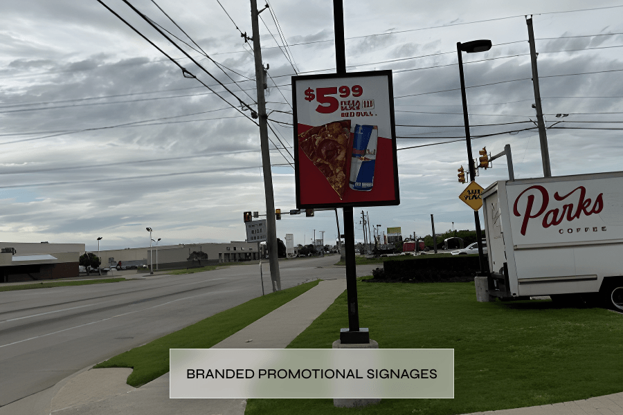 A promotional outdoor sign advertising pizza and a drink for $5.99, with a pizza slice and a can of soda depicted on the sign, in a commercial area with power lines, traffic lights, and buildings in the background.