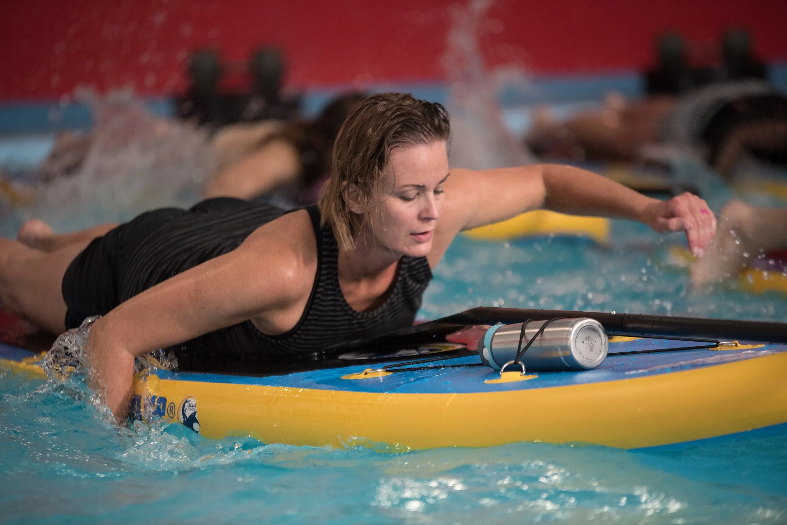 Woman doing a crawl exercise on a floating mat in a swimming pool, with other people also exercising in the background.